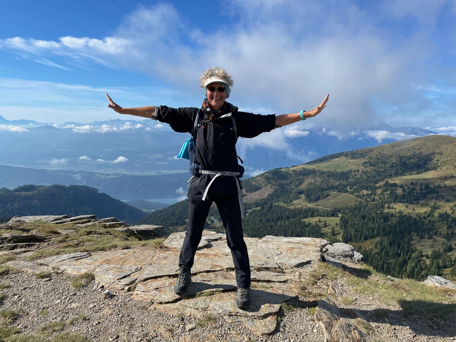 Mindtrail - Coaching und Mentoring - Eine Wanderin steht mit ausgestreckten Armen auf einem felsigen Berggipfel und genießt den malerischen Blick auf ferne Berge und einen See unter einem blauen Himmel mit weißen Wolken.